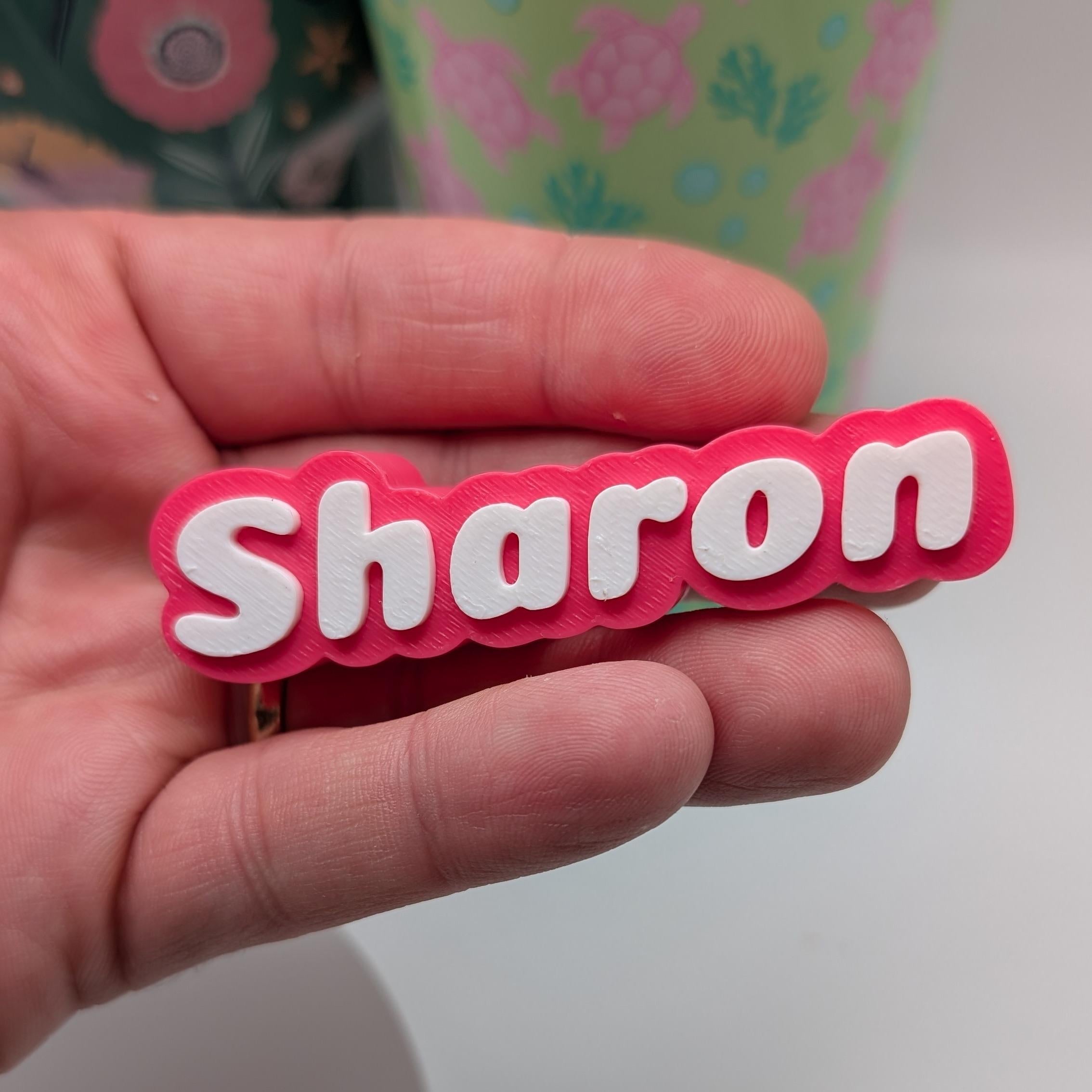Hand holding a pink and white 'Sharon' name tag against a light background